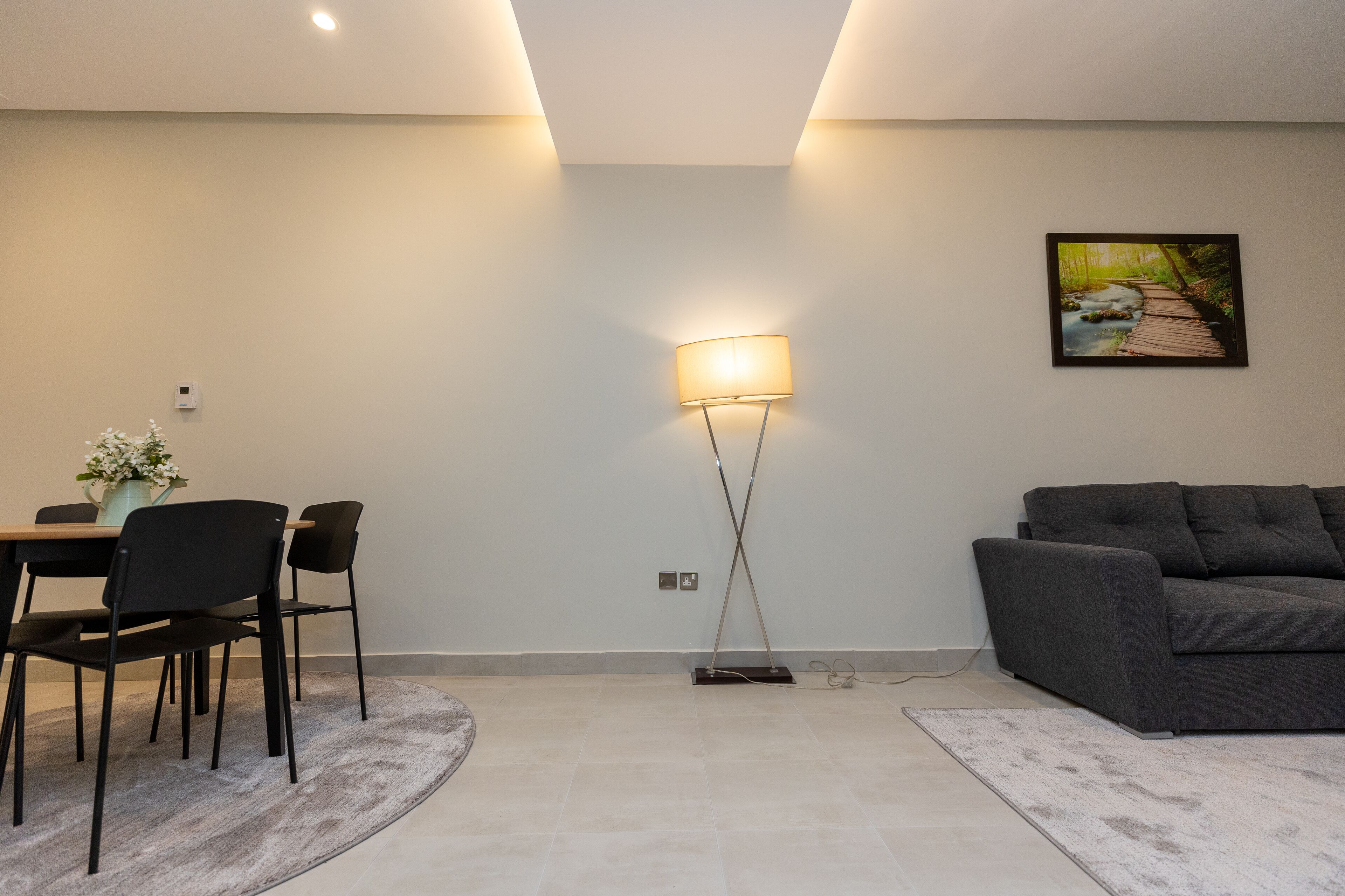 Een moderne woonkamer met een grijze bank, een eettafel met stoelen, een vloerlamp en een schilderij aan de muur.