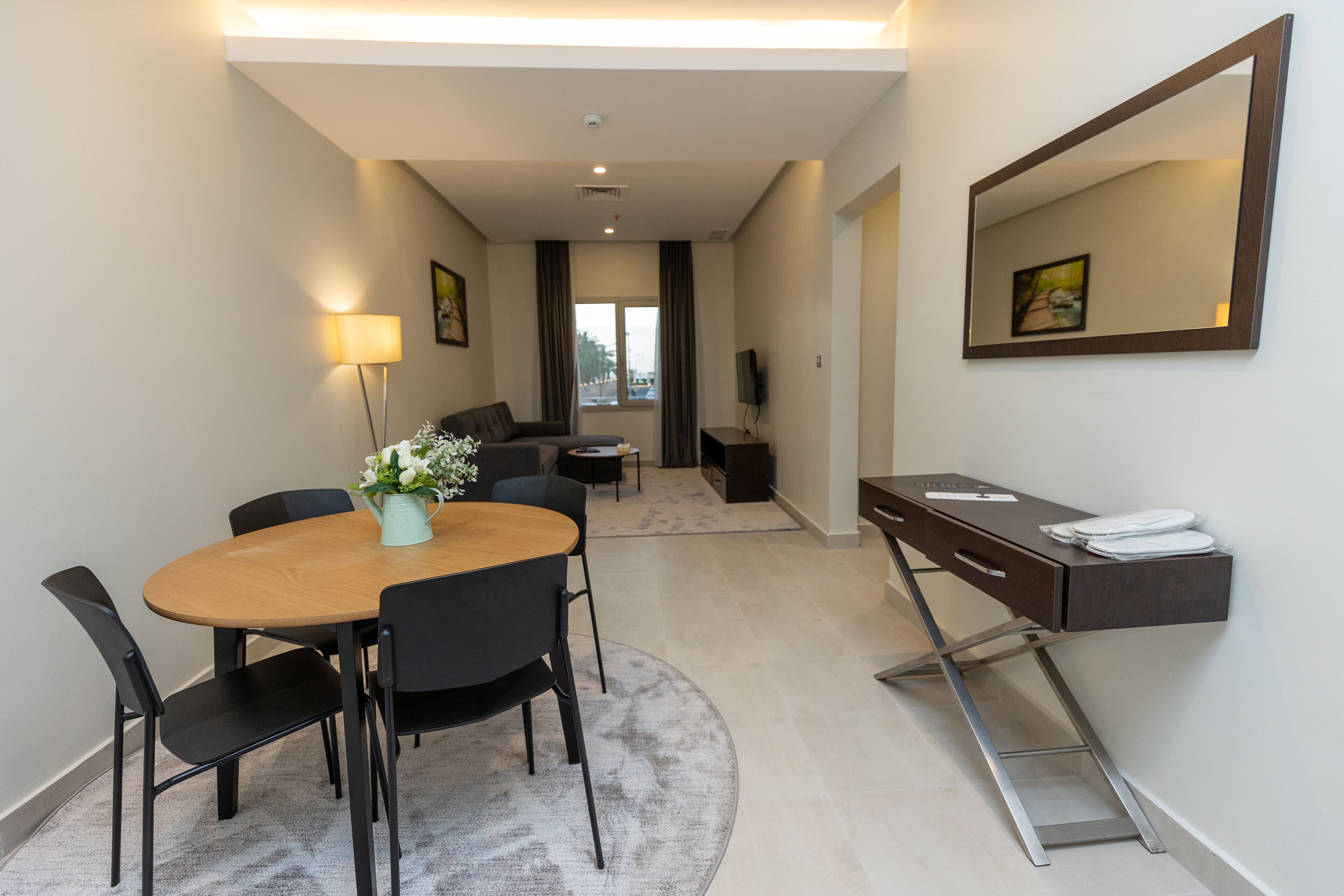 A modern hotel room with a dining area, a desk with a mirror, and a view of the outdoors.