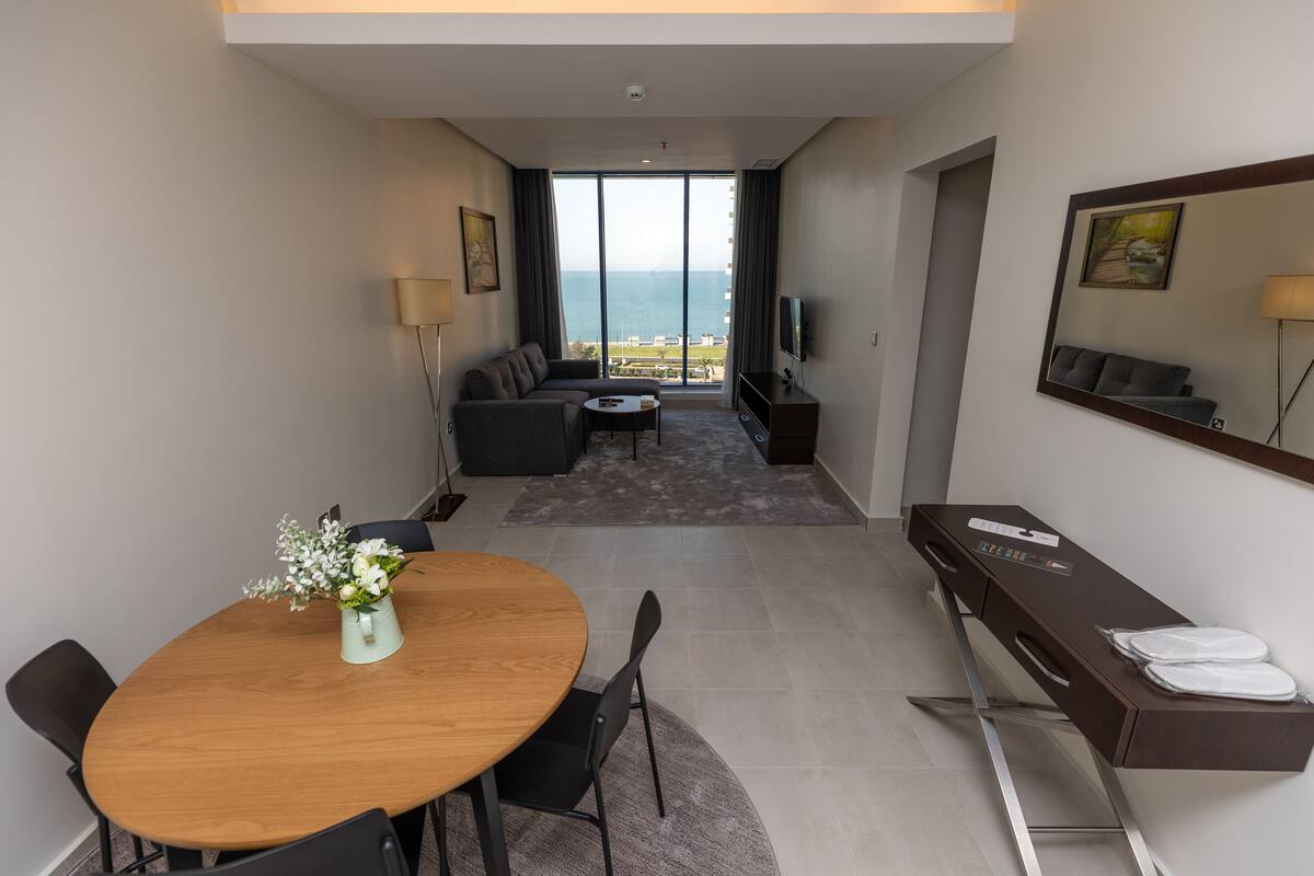 Quarto de hotel moderno com área de refeições, sofá, televisão e vista para o mar.