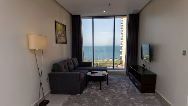 Un salon moderne avec un canapé, une table basse, un téléviseur et une grande fenêtre donnant sur la mer.