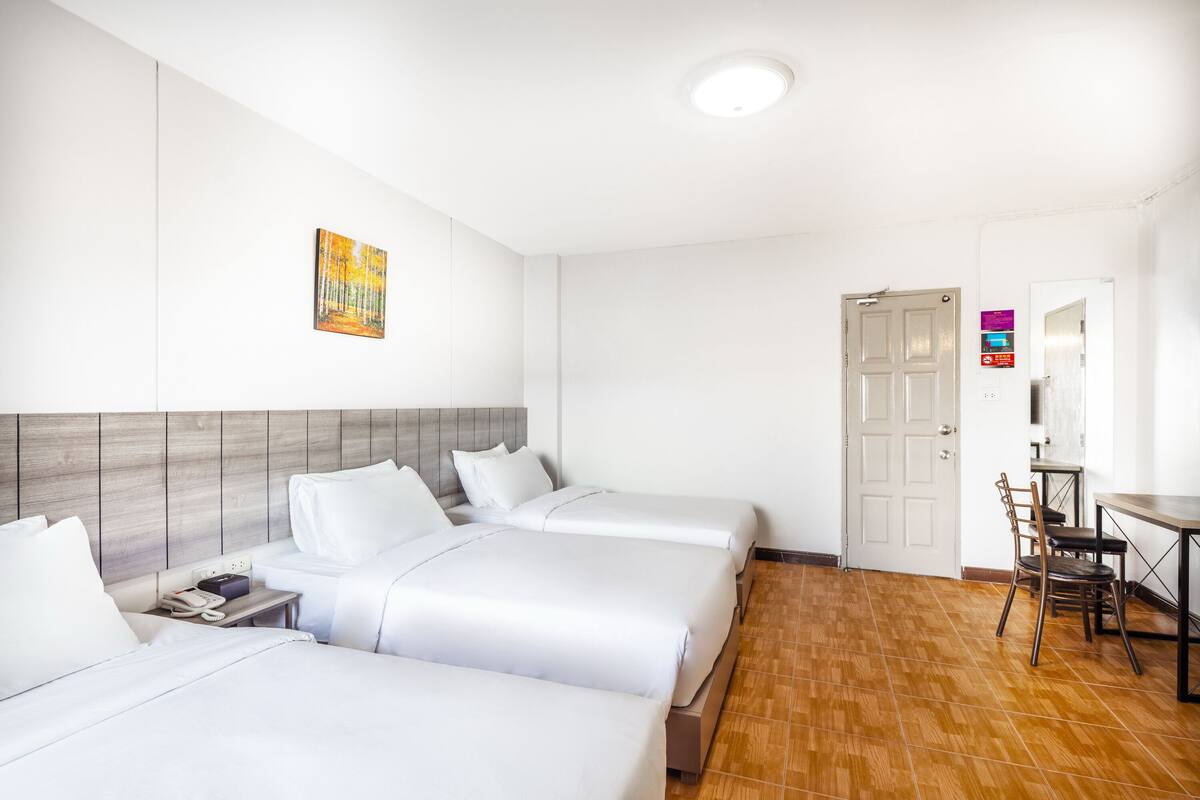 Hotellihuone, jossa on kolme sänkyä, puinen pääty, taulu seinällä, työpöytä tuolilla ja ovi.