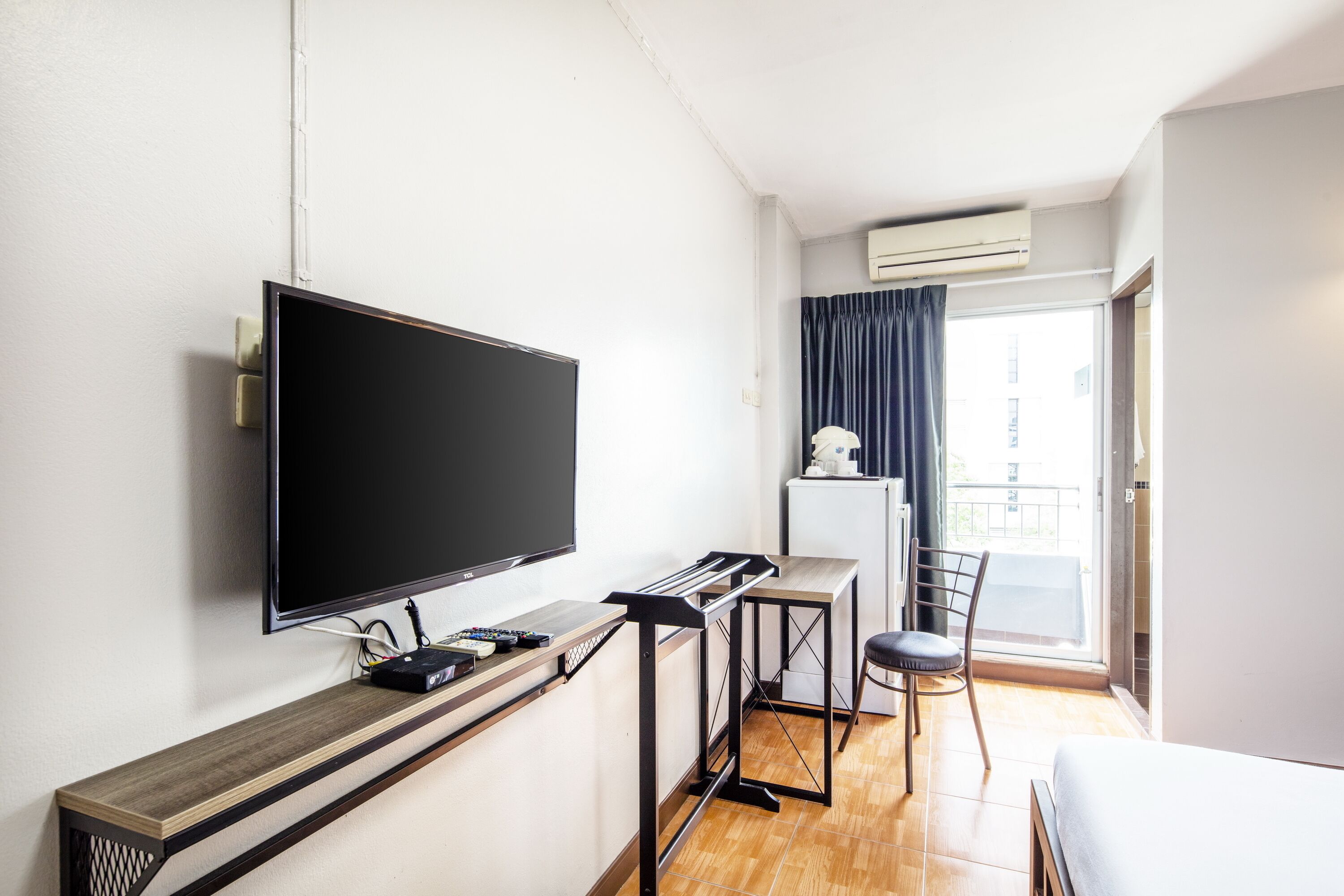 A hotel room with a flat-screen TV mounted on the wall, a small desk with a computer, a chair, and a bed.