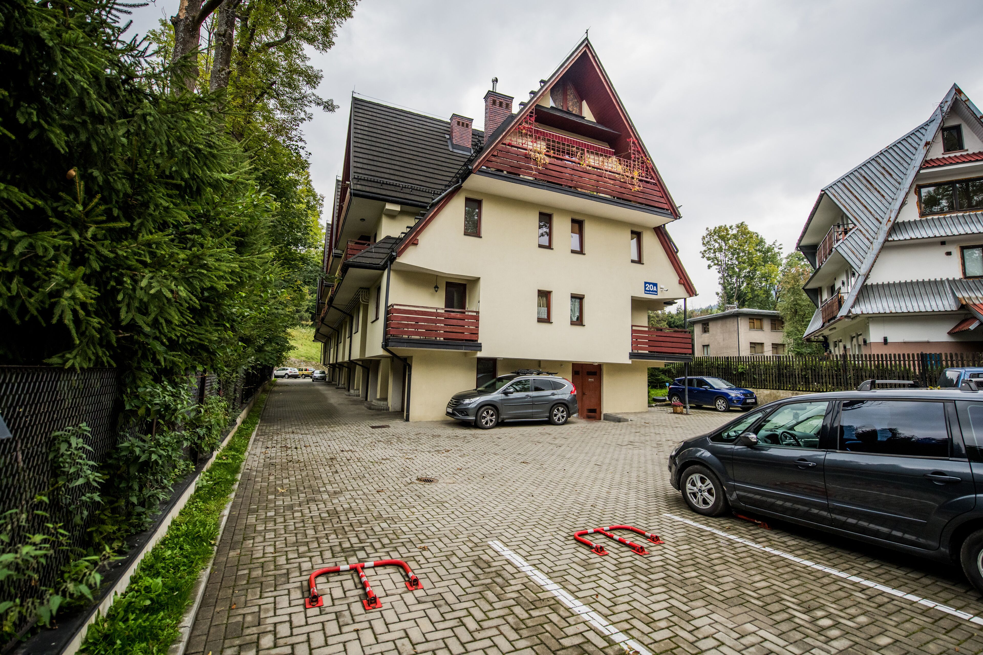 Um edifício residencial com telhado inclinado, varanda e entrada pavimentada com carros estacionados.