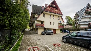 Um edifício residencial com telhado inclinado, varanda e entrada pavimentada com carros estacionados.