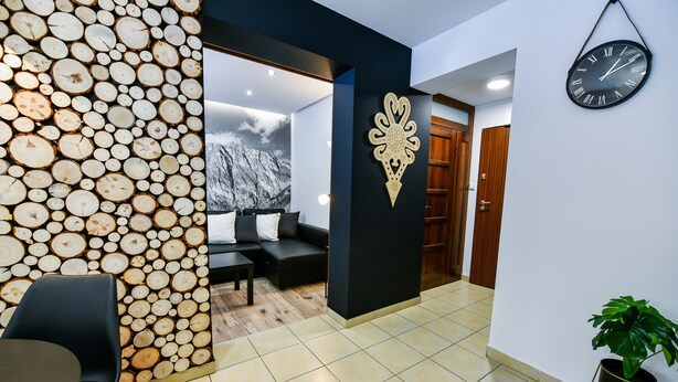 A modern interior with a wooden log wall, a black sofa, a dining area, and a clock on the wall. - Rentplanet - Bulwary Słowackiego (Zakopane)