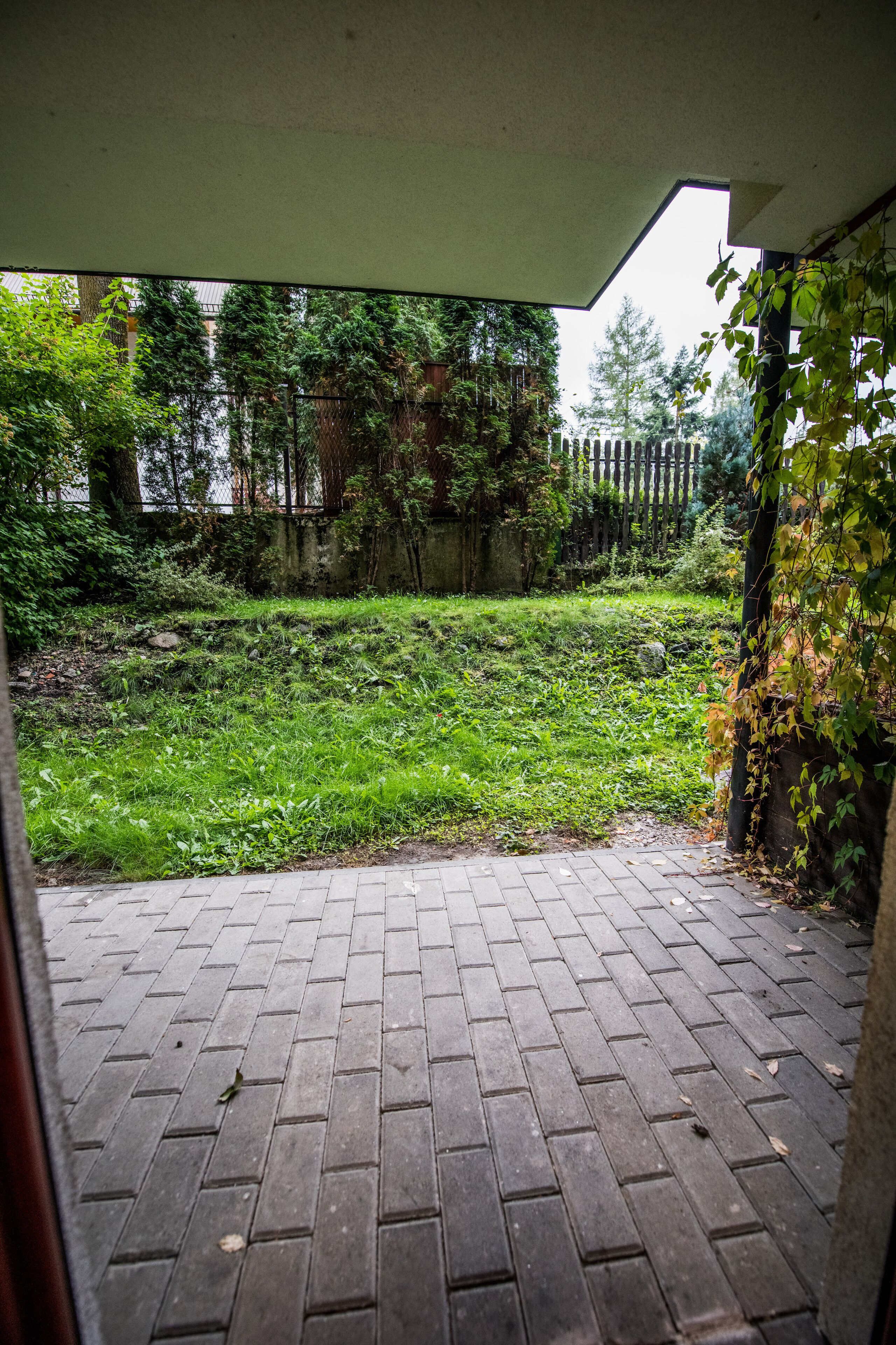 Uma área de pátio externa com piso de tijolos e vista para uma área gramada com árvores e uma cerca ao fundo.