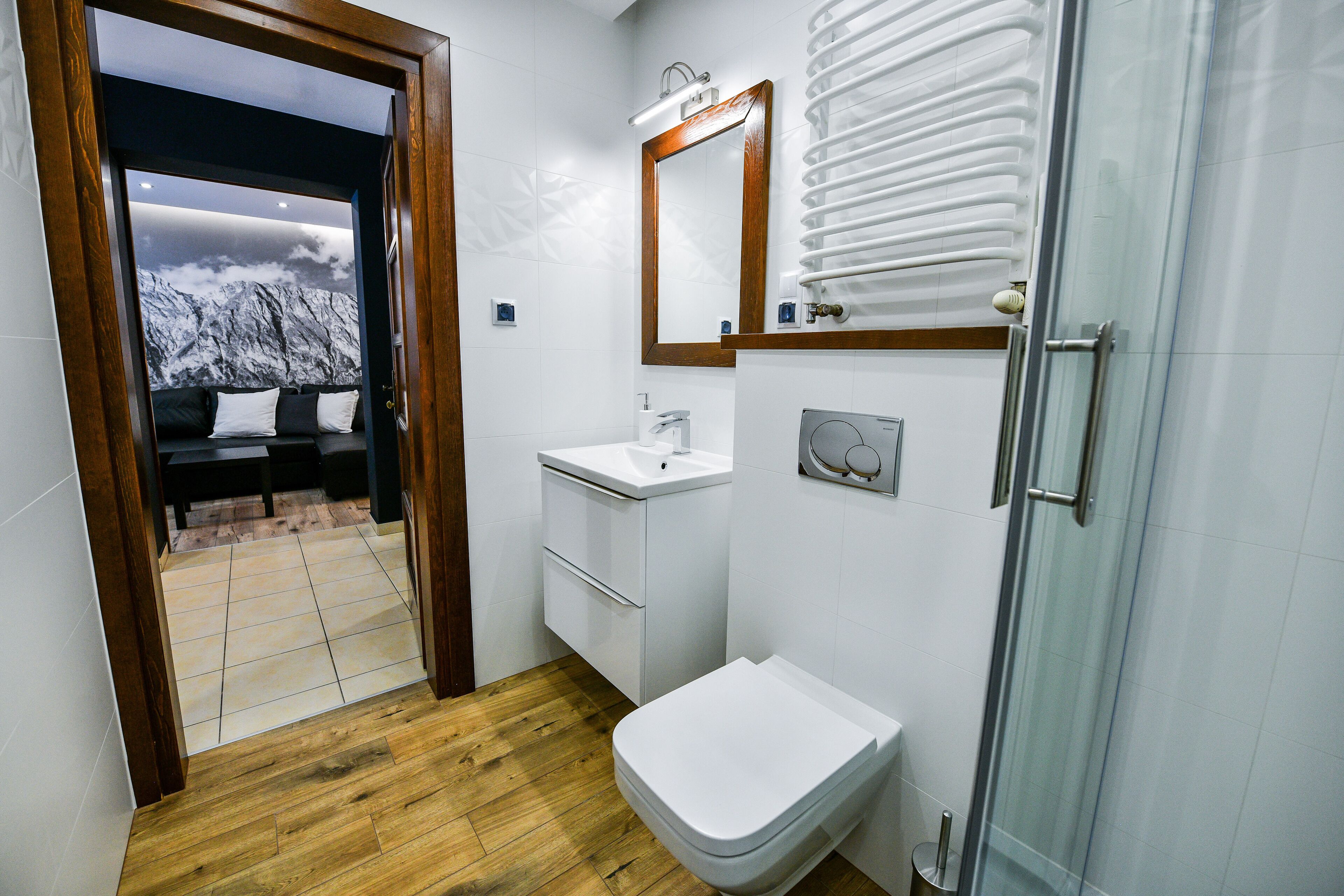 Un bany amb sòl de fusta, elements blancs i una cabina de dutxa de vidre, i vista a una muntanya nevada a través d'una porta.