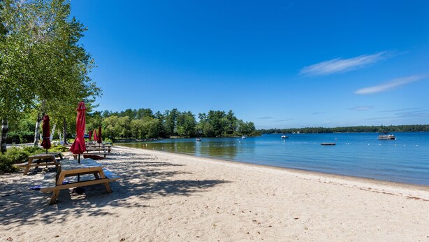 Ein Strand mit Holzliegen und roten Sonnenschirmen, ein sandiger Uferstreifen und ein ruhiger See mit Booten.