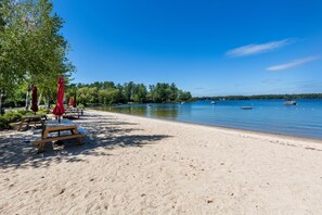 Une plage avec des bancs en bois et des parasols rouges, un rivage sablonneux et un lac paisible où flottent des bateaux.
