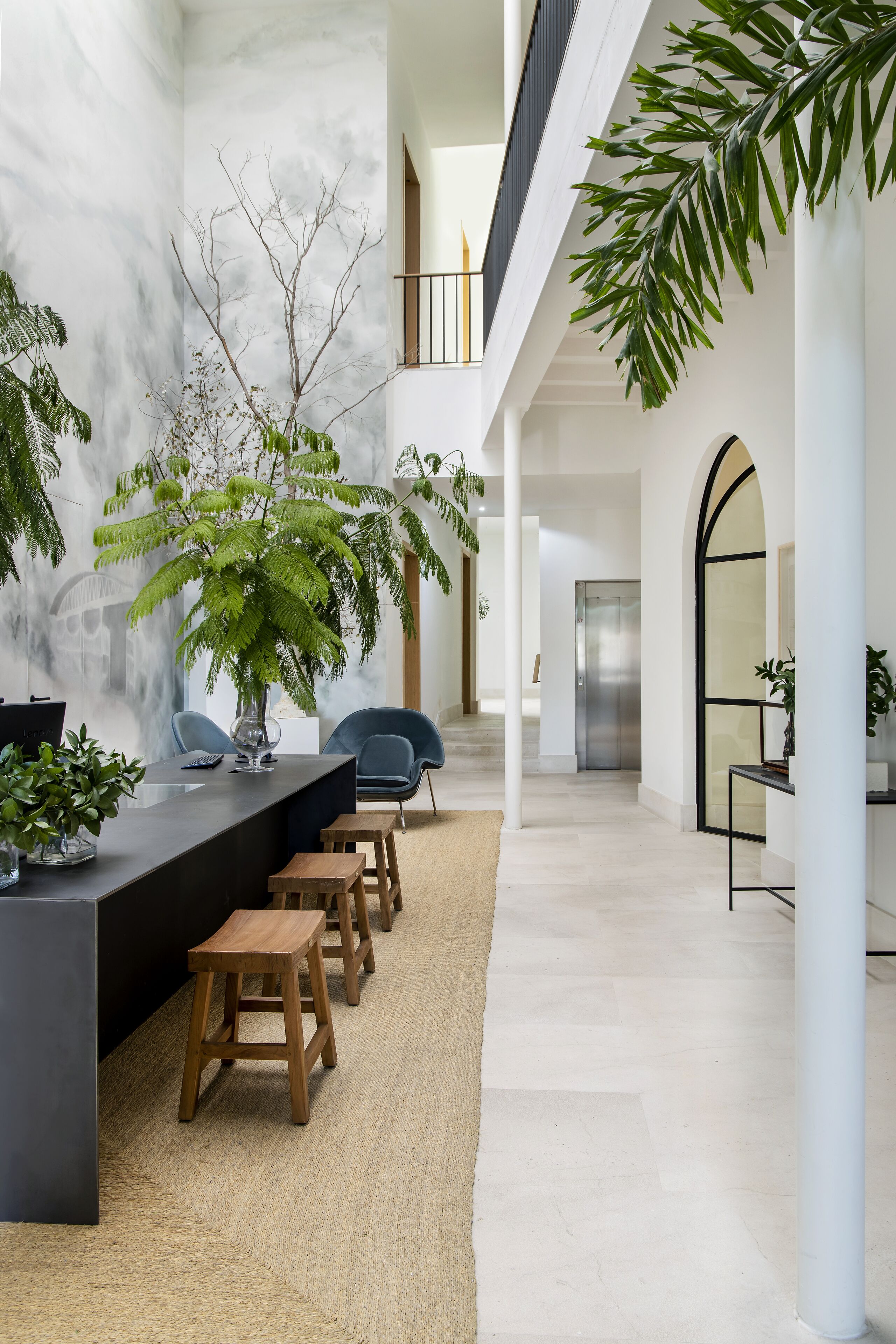 Decoração moderna com balcão de recepção, banquetas de madeira e grandes plantas verdes.