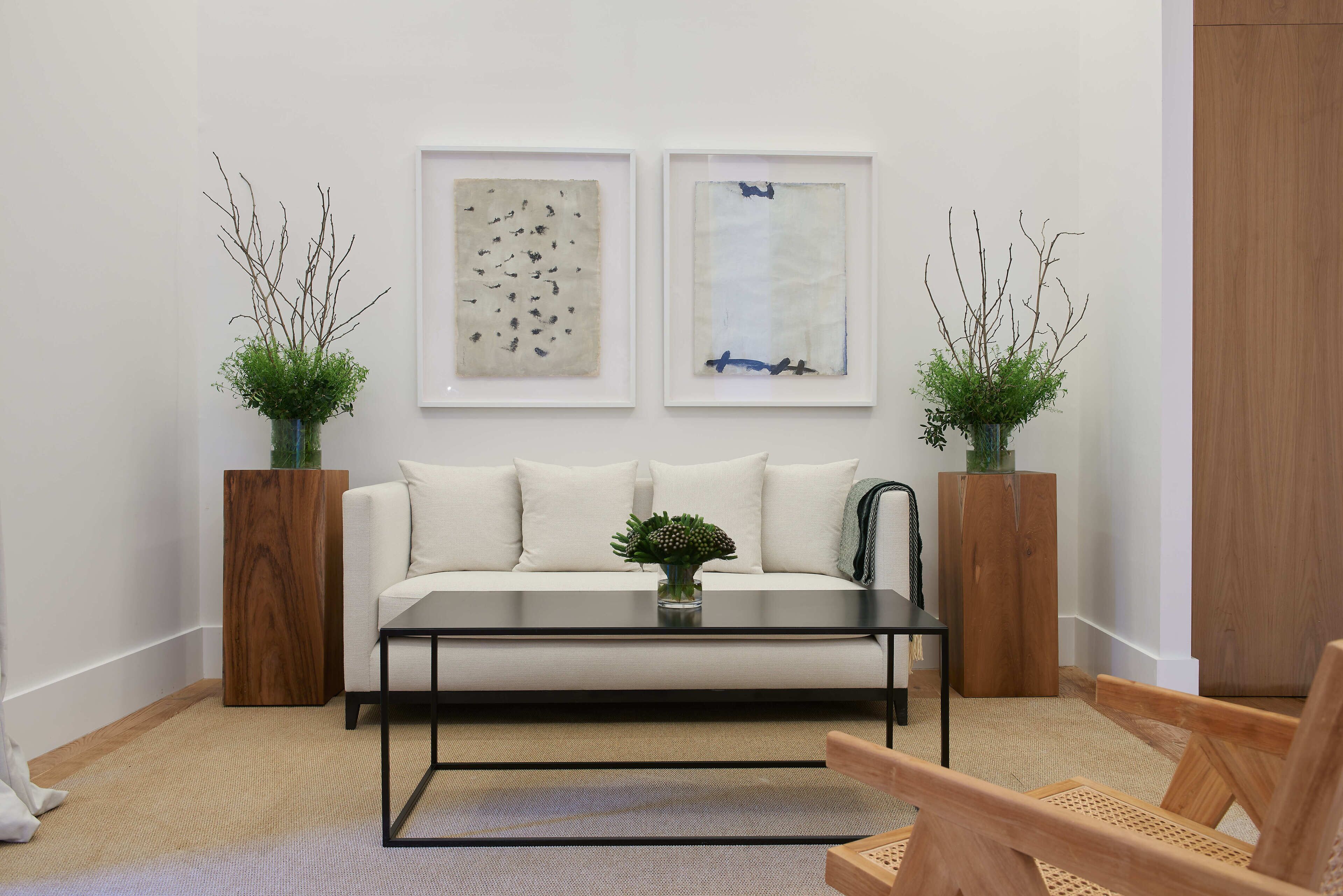 A modern living room with a white sofa, a glass coffee table, and two abstract paintings on the wall. There are two wooden side tables with plants, and a wooden chair in the foreground.
