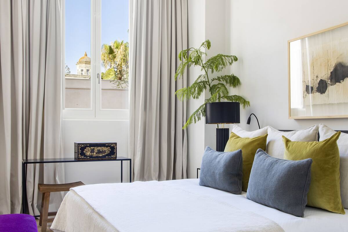 Une chambre à coucher avec un lit, une table de chevet, une lampe, une plante et une fenêtre avec des rideaux.
