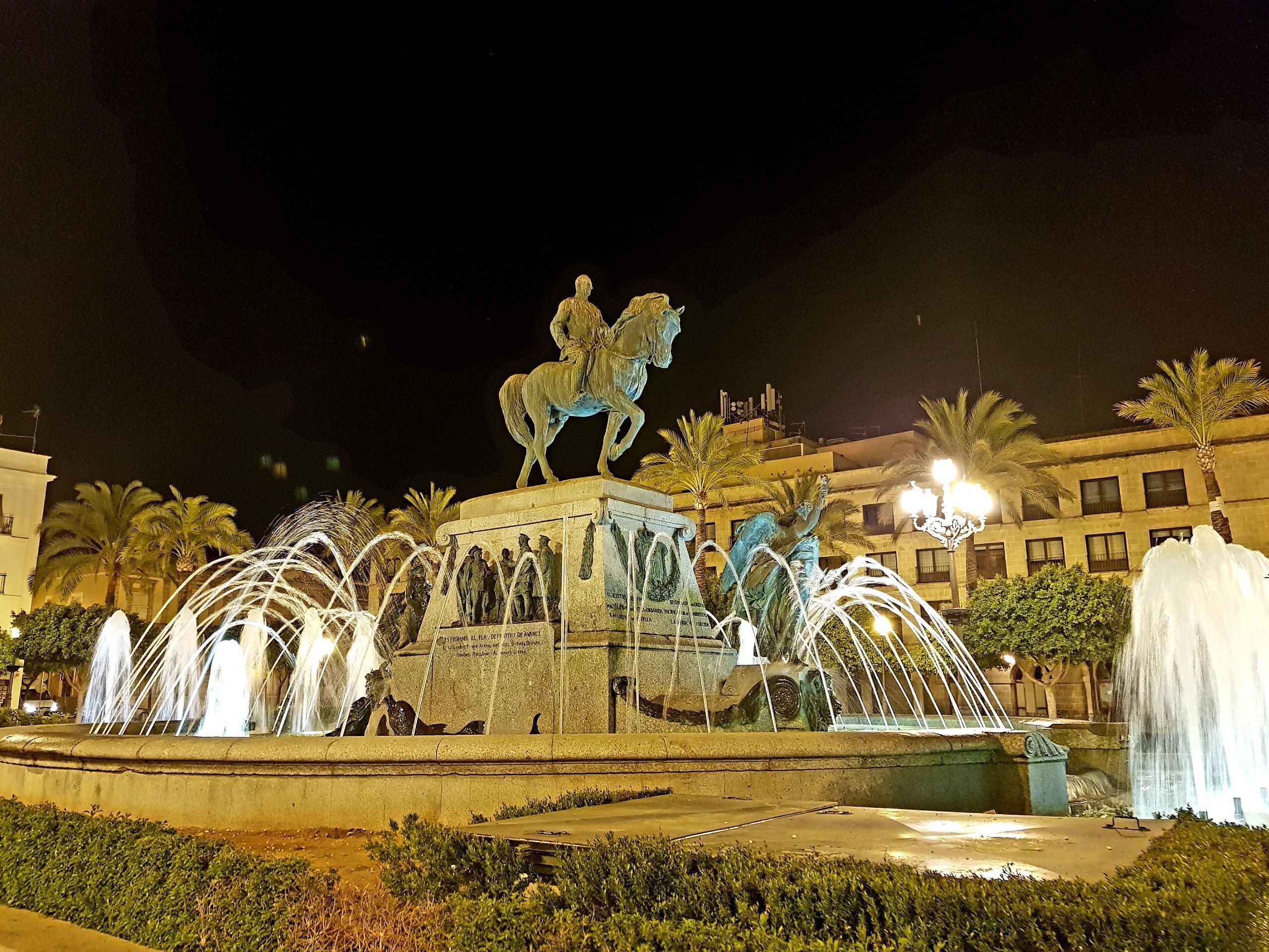 A nighttime fountain with a statue of a man on a horse, surrounded by water jets and palm trees.