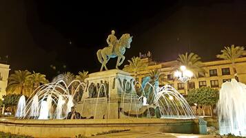 A nighttime fountain with a statue of a man on a horse, surrounded by water jets and palm trees.