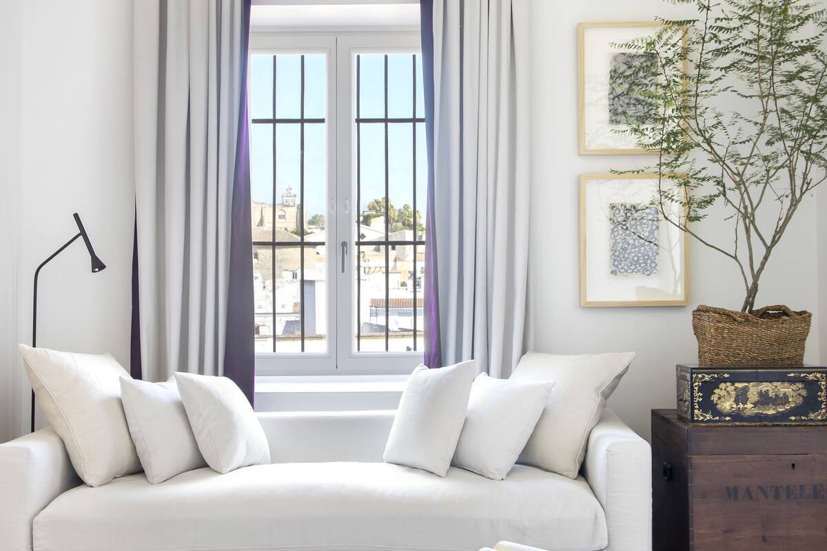 Un salon moderne avec un canapé blanc, un pouf violet et une vue dégagée sur l’extérieur grâce à une grande fenêtre agrémentée de rideaux.