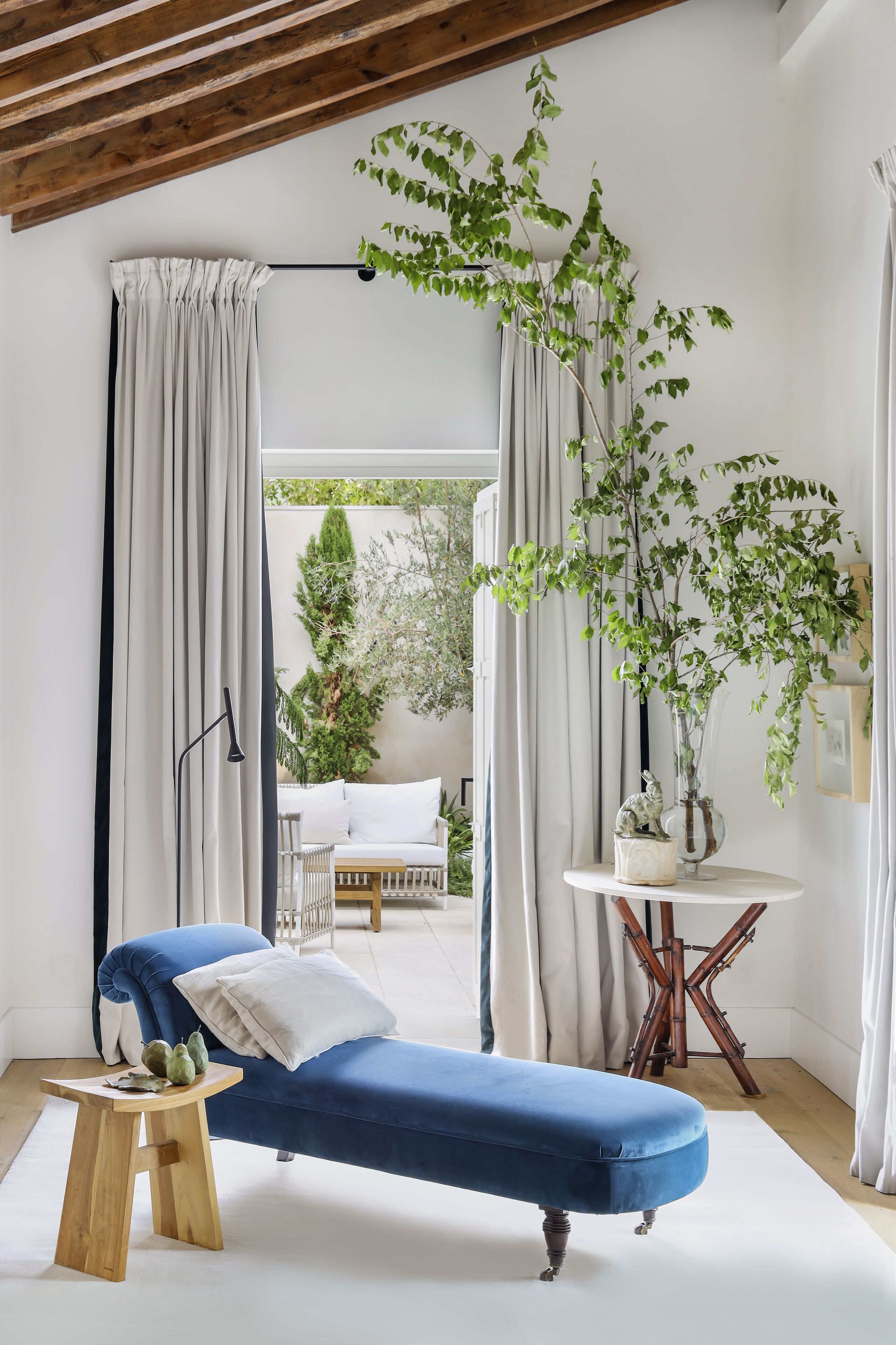 Una habitación con un sofá azul, un taburete de madera, una mesa redonda con un florero y una planta grande.