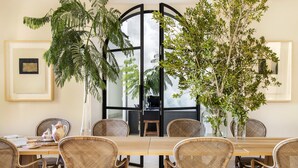 Uma área de refeições com uma mesa de madeira comprida, cadeiras de vime e plantas em vasos.