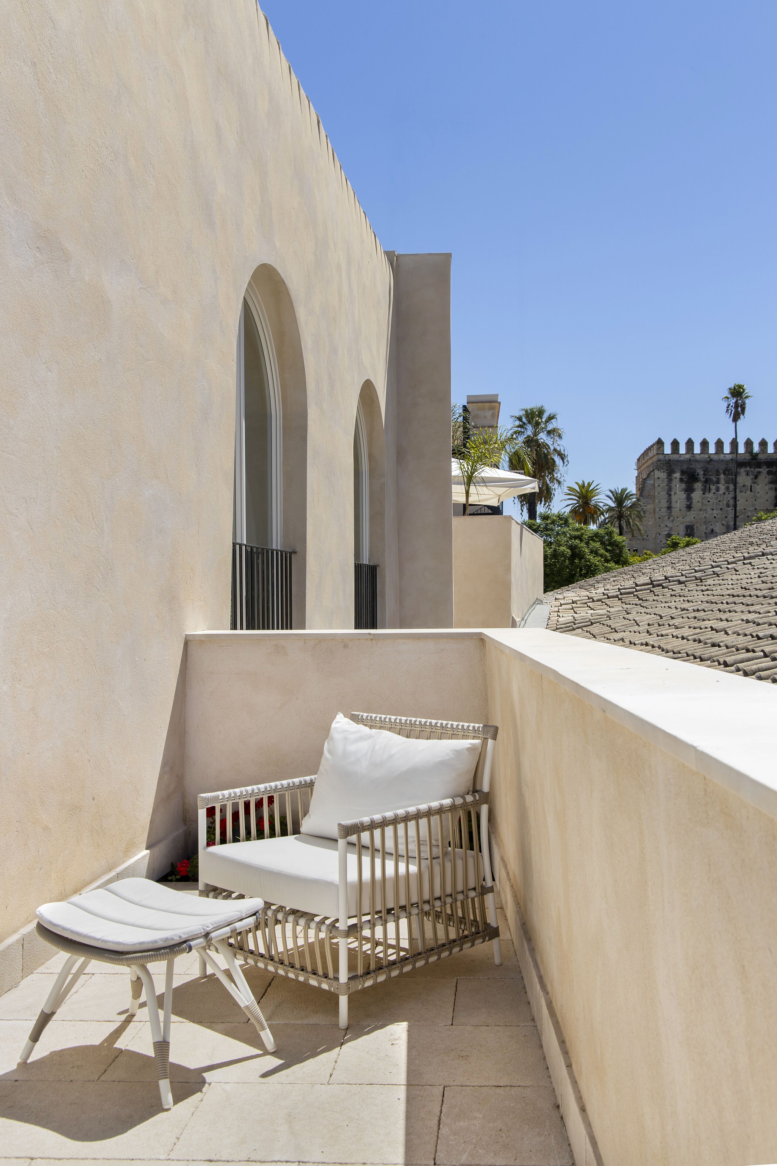A sunlit outdoor seating area with a white cushioned chair and a small table, adjacent to a building with arched windows and a stone wall.