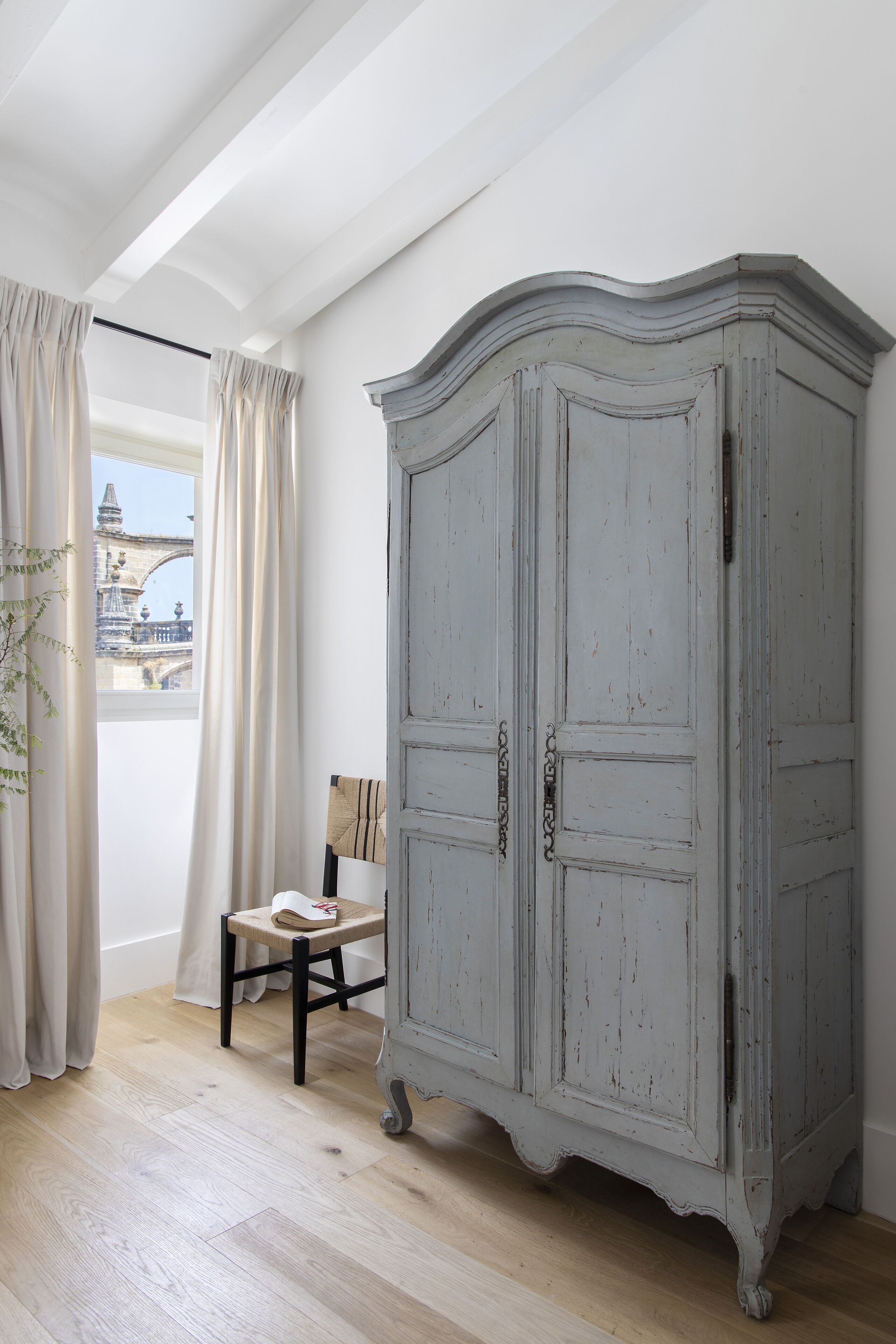 Una habitación con suelo de madera clara, techo blanco con tragaluces, un armario gris, una silla y vistas a un puente a través de una ventana con cortinas.