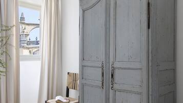 Una habitación con suelo de madera clara, techo blanco con tragaluces, un armario gris, una silla y vistas a un puente a través de una ventana con cortinas.