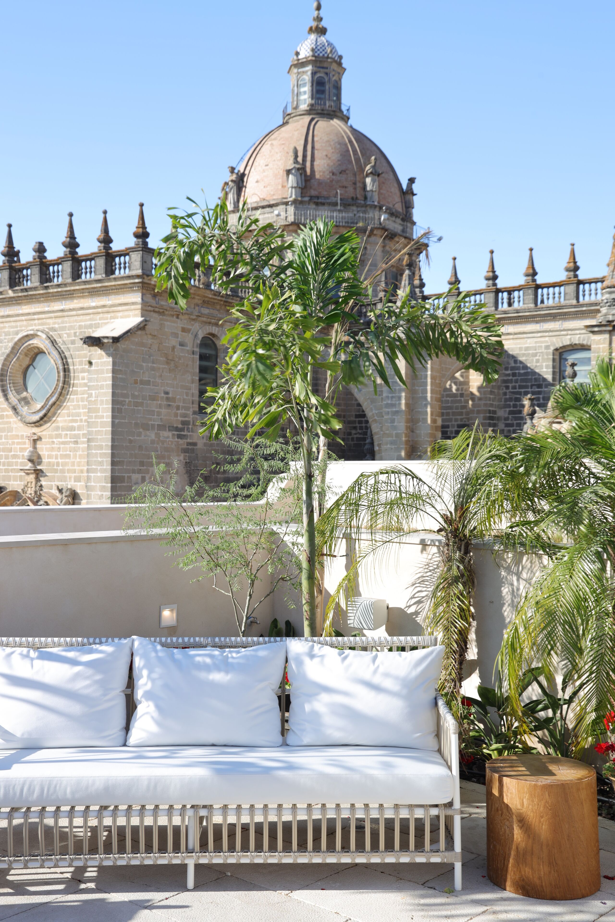 Un sofá blanco al aire libre con cojines, un taburete de madera y una palmera frente a un edificio histórico con una cúpula y ventanas arqueadas.