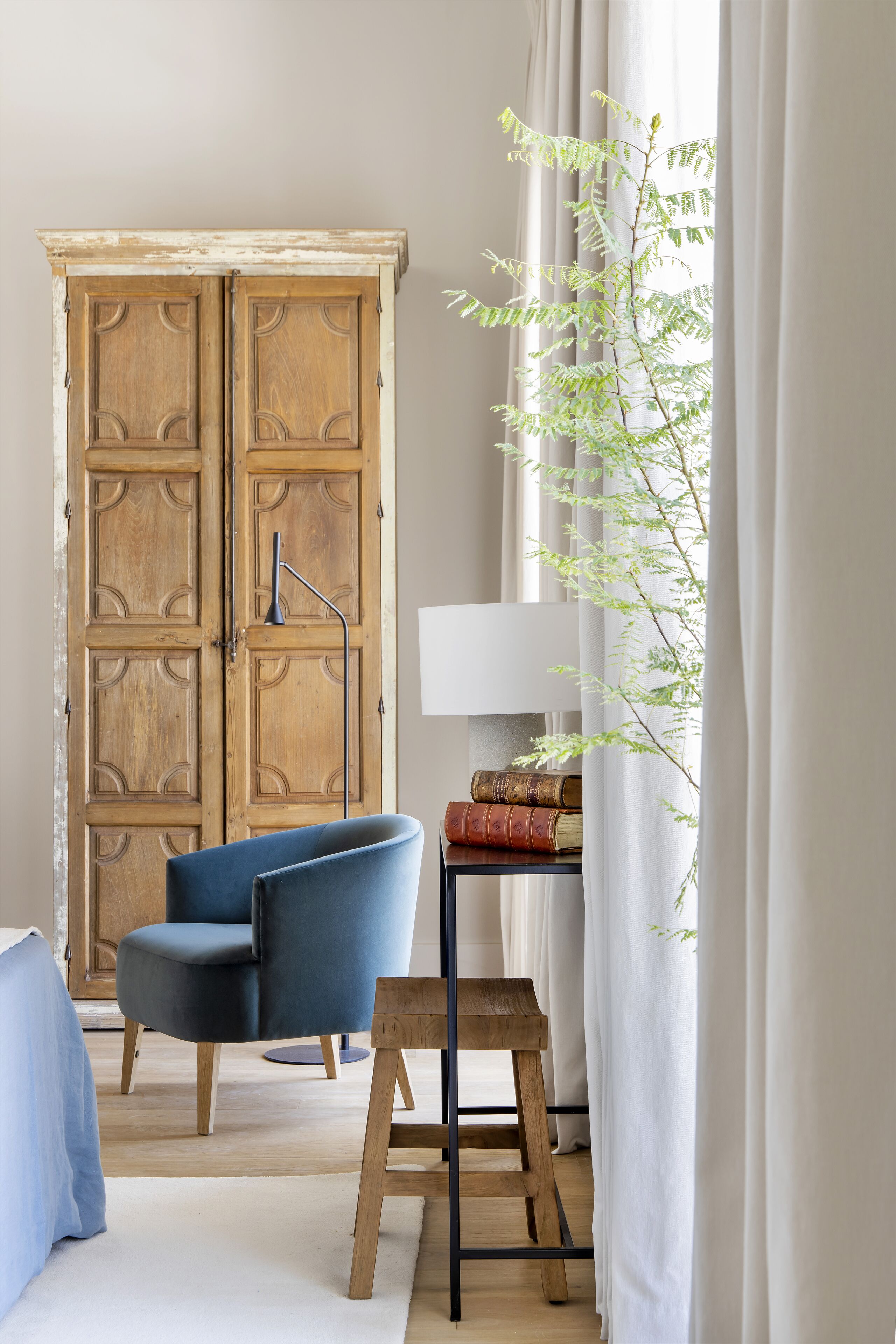 A room with a wooden wardrobe, a blue chair, a small wooden stool, and a floor lamp.