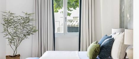 A bedroom with a bed, a bedside table, a lamp, a window with curtains, and a potted plant.