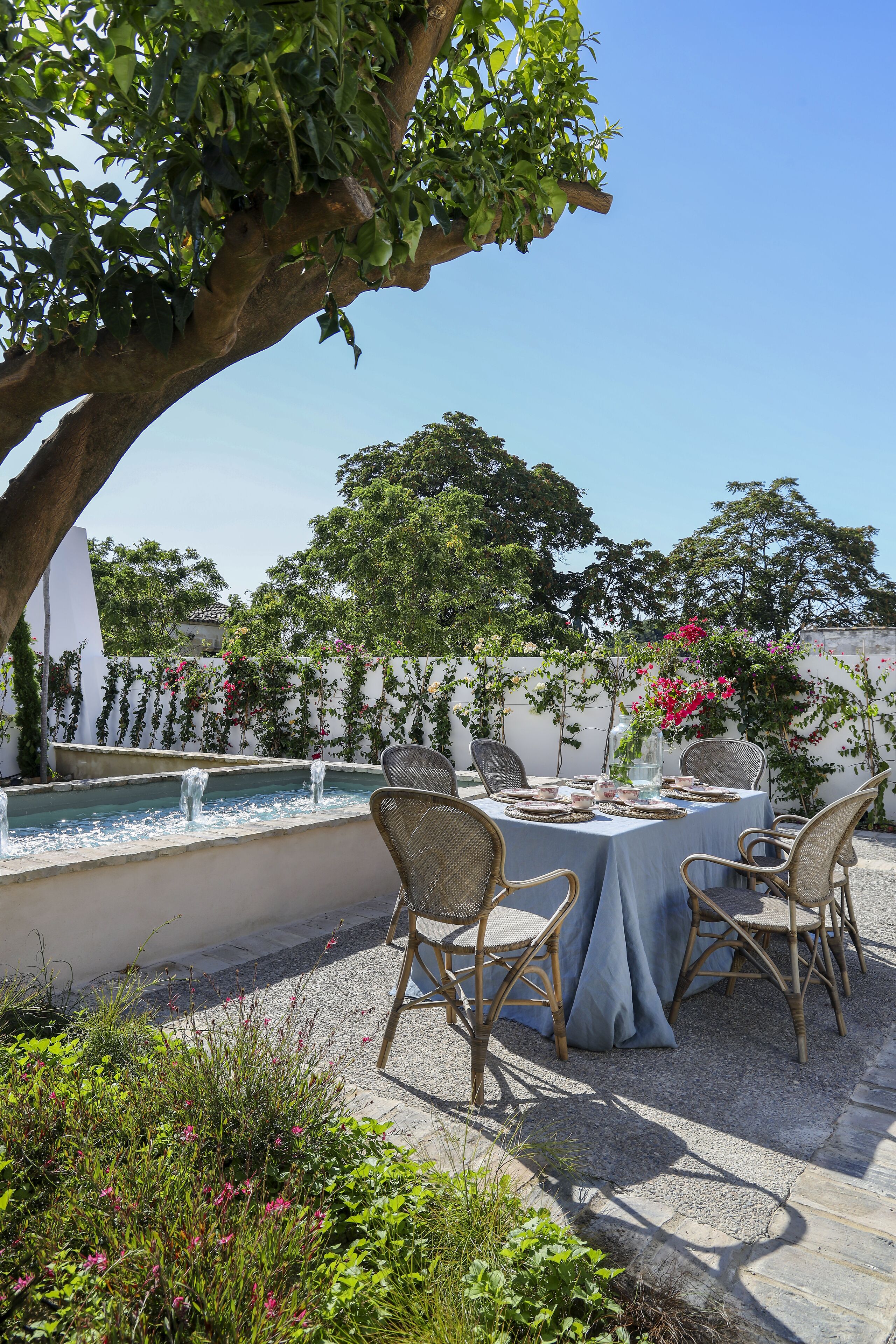 An outdoor patio with a round table, four wicker chairs, a small fountain, and lush greenery.