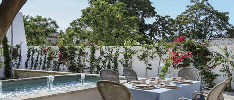 An outdoor patio with a round table, four wicker chairs, a small fountain, and lush greenery.