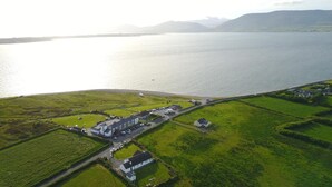 An aerial view of a coastal area with a large body of water, green fields, and a building complex with parking. - Jacks Coastguard Cottage (Killorglin)