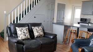A living room with a black leather sofa, two patterned throw pillows, a wooden coffee table, and a kitchen area with a microwave and wooden stools. - Jacks Coastguard Cottage (Killorglin)