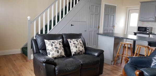A living room with a black leather sofa, two patterned throw pillows, a wooden coffee table, and a kitchen area with a microwave and wooden stools. - Jacks Coastguard Cottage (Killorglin)
