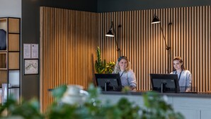 A reception desk with two computer monitors, a plant, and a wooden panel wall. - Comfort Hotel Copenhagen Airport (Kastrup)