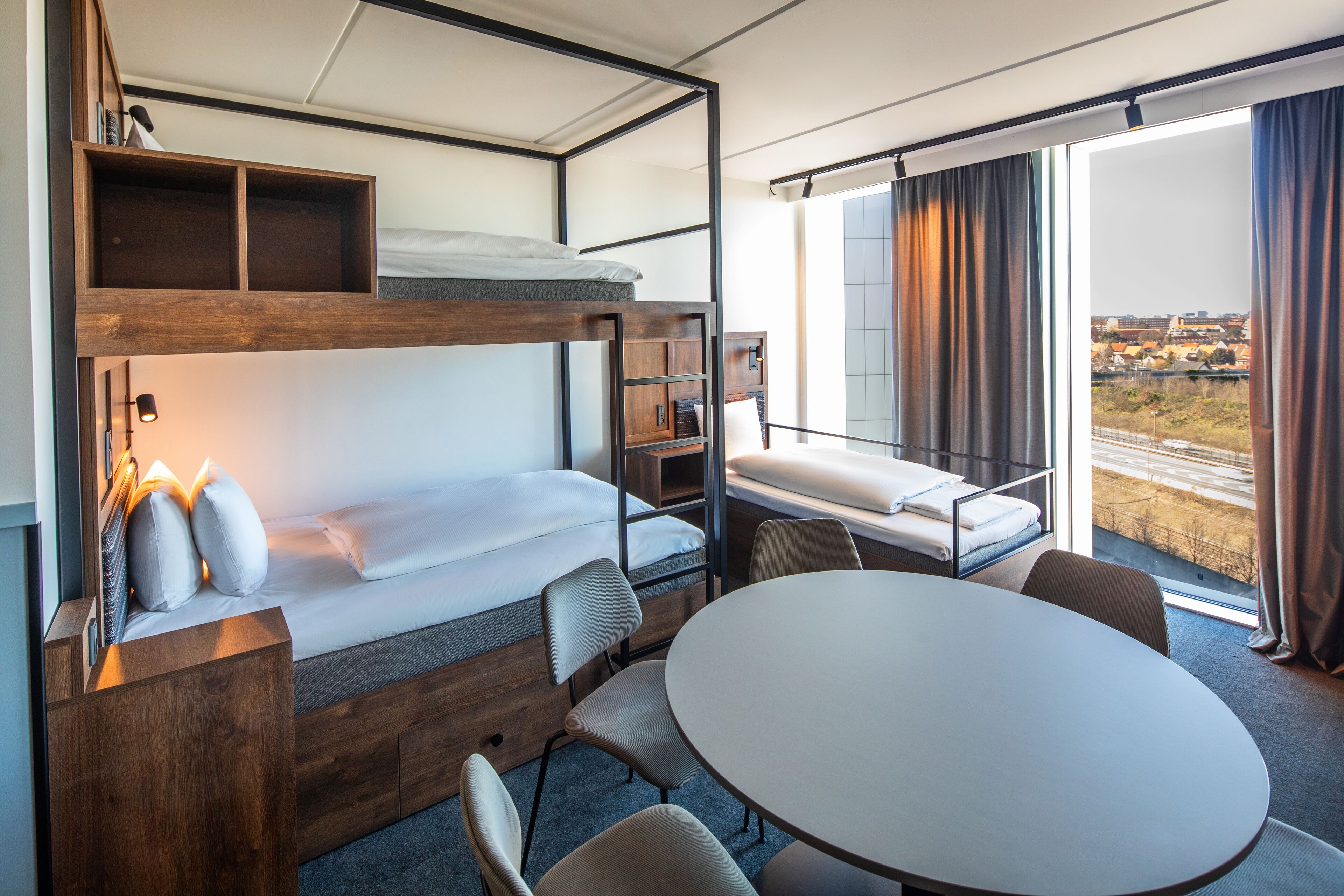 A compact hotel room with bunk beds, a small desk, a round table, and a view of the outside.