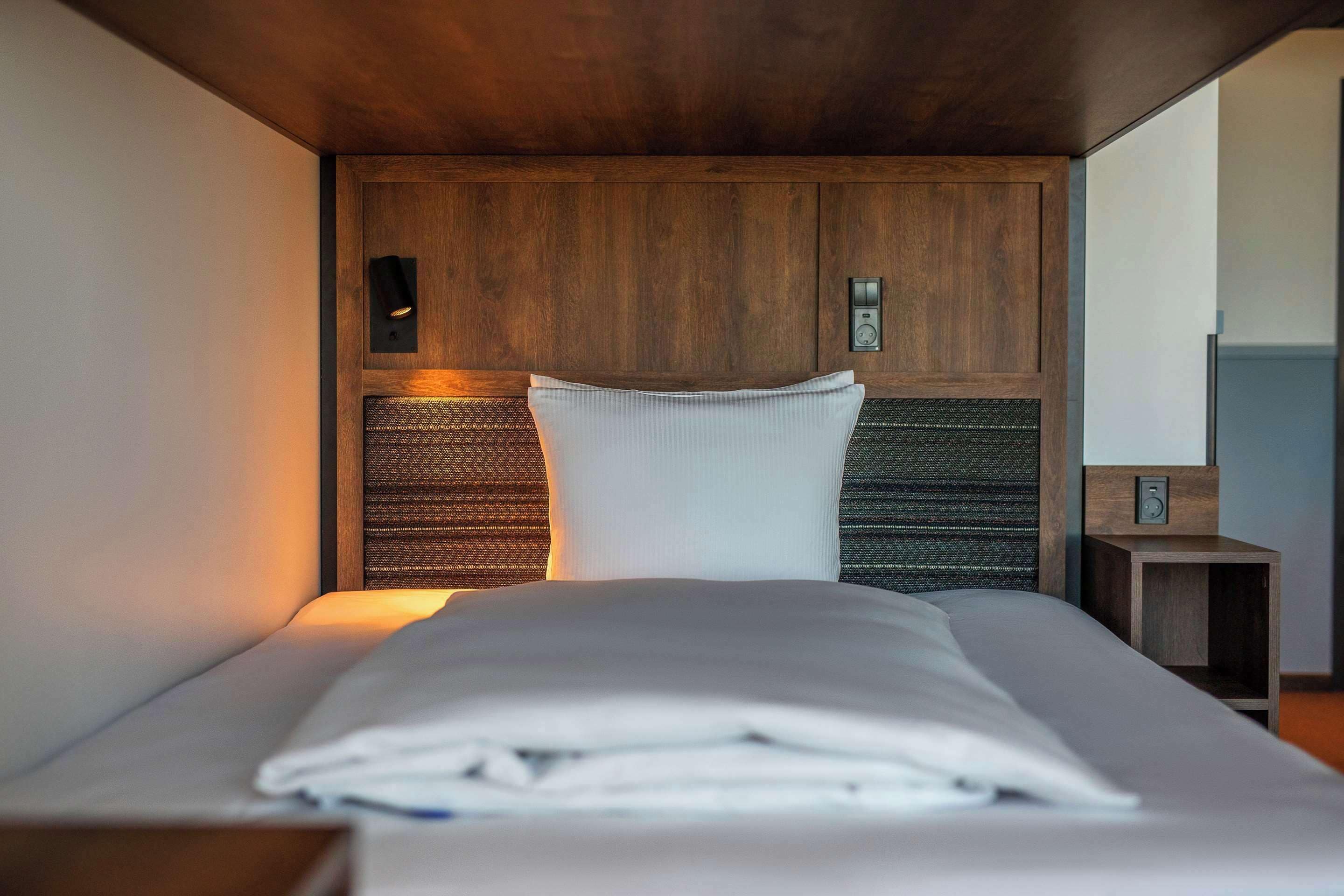 A neatly made bed with a white comforter and pillows, a wooden headboard, and a small bedside table with a lamp.