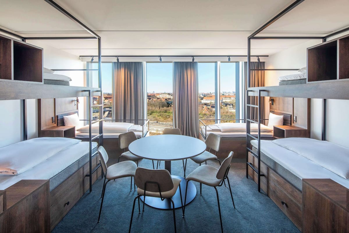 A modern hotel room with bunk beds, a round table, and chairs, large windows with curtains, and a view of the city.