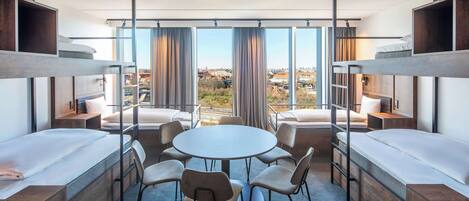 A modern hotel room with bunk beds, a round table, and chairs, large windows with curtains, and a view of the city.