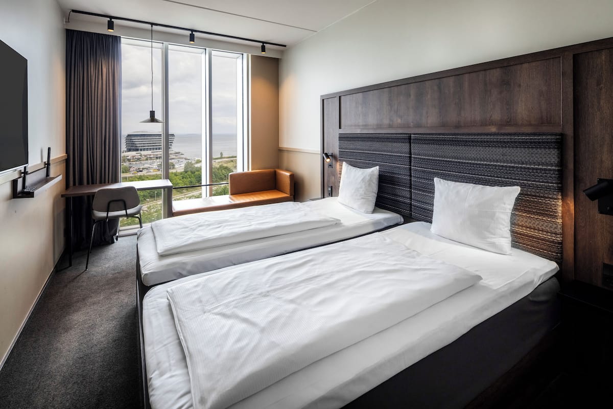 A hotel room with two beds, a desk, a chair, and a large window offering a city view.