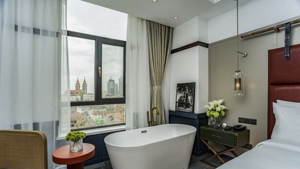 A modern hotel room with a large bathtub, a city view, a small table with a vase, and a bed with a headboard. - Manxin Hotel Qingdao Zhanqiao (Qingdao)