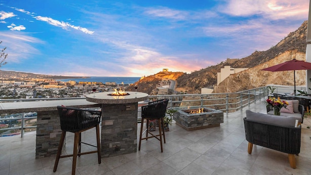 A terrace with a stone fire pit, two bar stools, a cushioned chair, and a table with a red umbrella. - Luxury PH Cascadas in Cabo Central Location, Ocean Views, 5-Star Service (Pedregal)