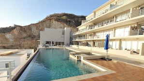A swimming pool with a wooden deck, a modern building with balconies, and a hillside in the background. - Luxury PH Cascadas in Cabo Central Location, Ocean Views, 5-Star Service (Pedregal)
