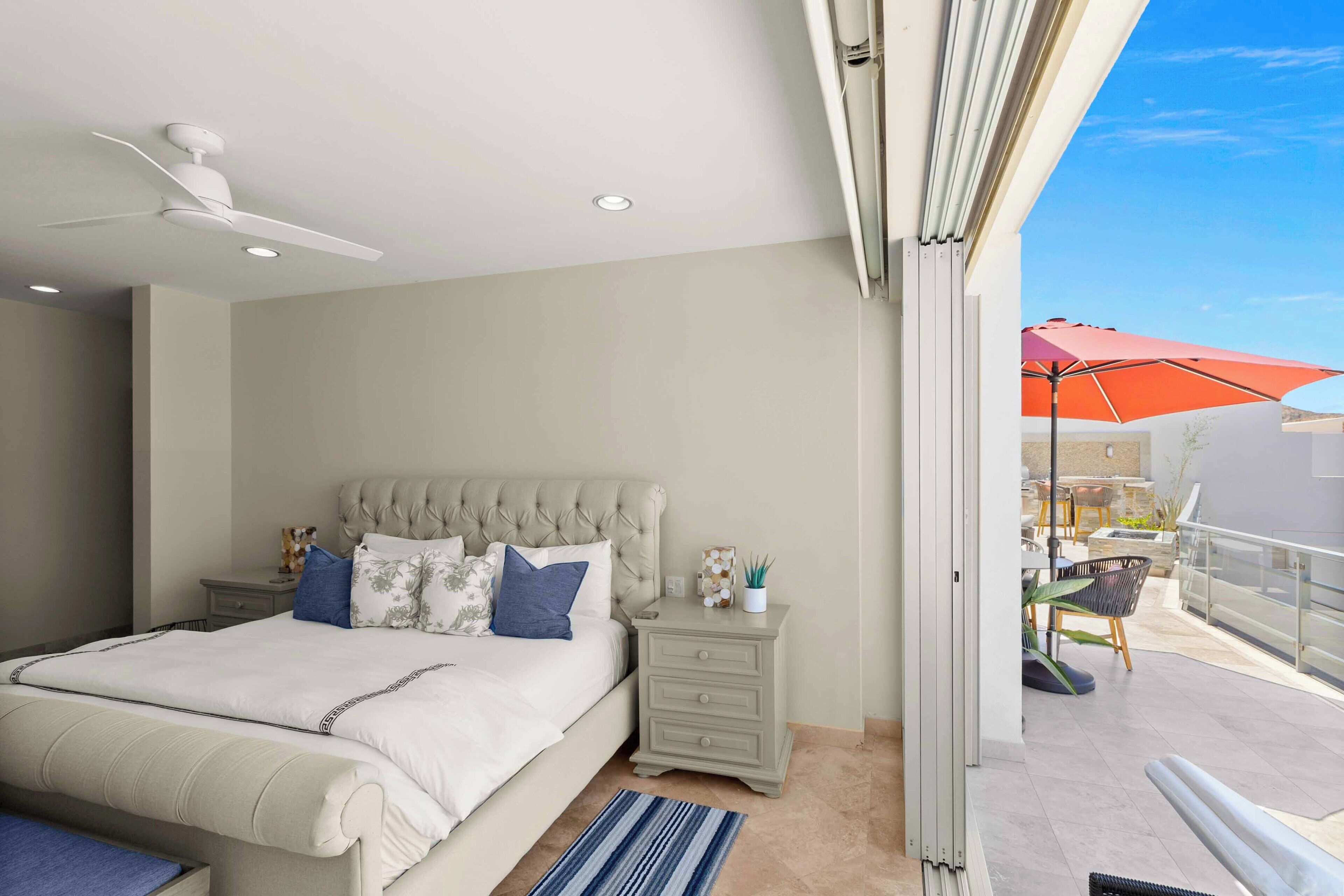 A bedroom with a large bed, a nightstand, a ceiling fan, and a balcony with outdoor furniture.