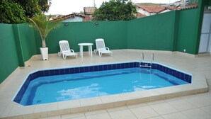 Ein rechteckiger Swimmingpool mit blauen Fliesen und einem weißen Rand. Zwei weiße Liegestühle und ein kleiner weißer Tisch mit einem Topf stehen in der Nähe.