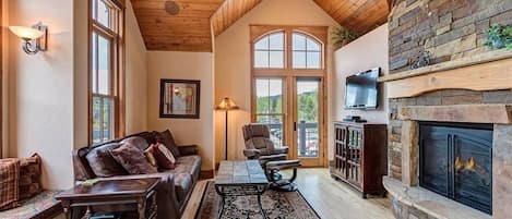 A cozy living room with a fireplace, wooden ceiling, and large windows.