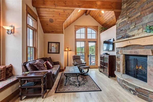 A cozy living room with a fireplace, wooden ceiling, and large windows.