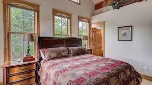 A bedroom with a large bed, wooden headboard, and multiple windows. There is a dresser with drawers and a lamp on it.