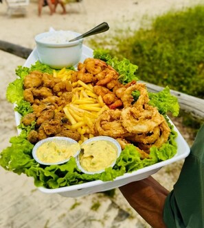 Una bandeja de mariscos fritos, incluyendo camarones, calamares y aros de calamar, acompañada de papas fritas y dos salsas para mojar, servida sobre una cama de lechuga.