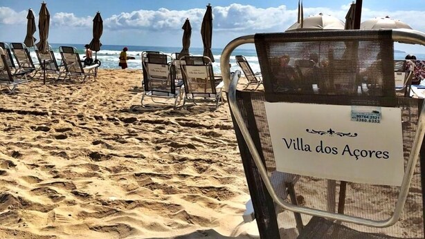 Una playa con filas de tumbonas y sombrillas, un letrero que dice "Villa dos Açores", y un cielo despejado.