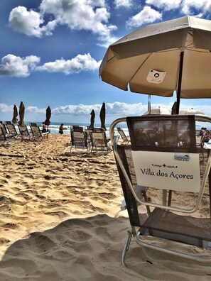 Une plage bordée de rangées de transats et de parasols, une pancarte indiquant « Villa dos Açores », et un ciel dégagé.