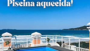 Piscina azul cristalina con cubierta de madera y barandillas blancas, con vistas a una playa y al océano.